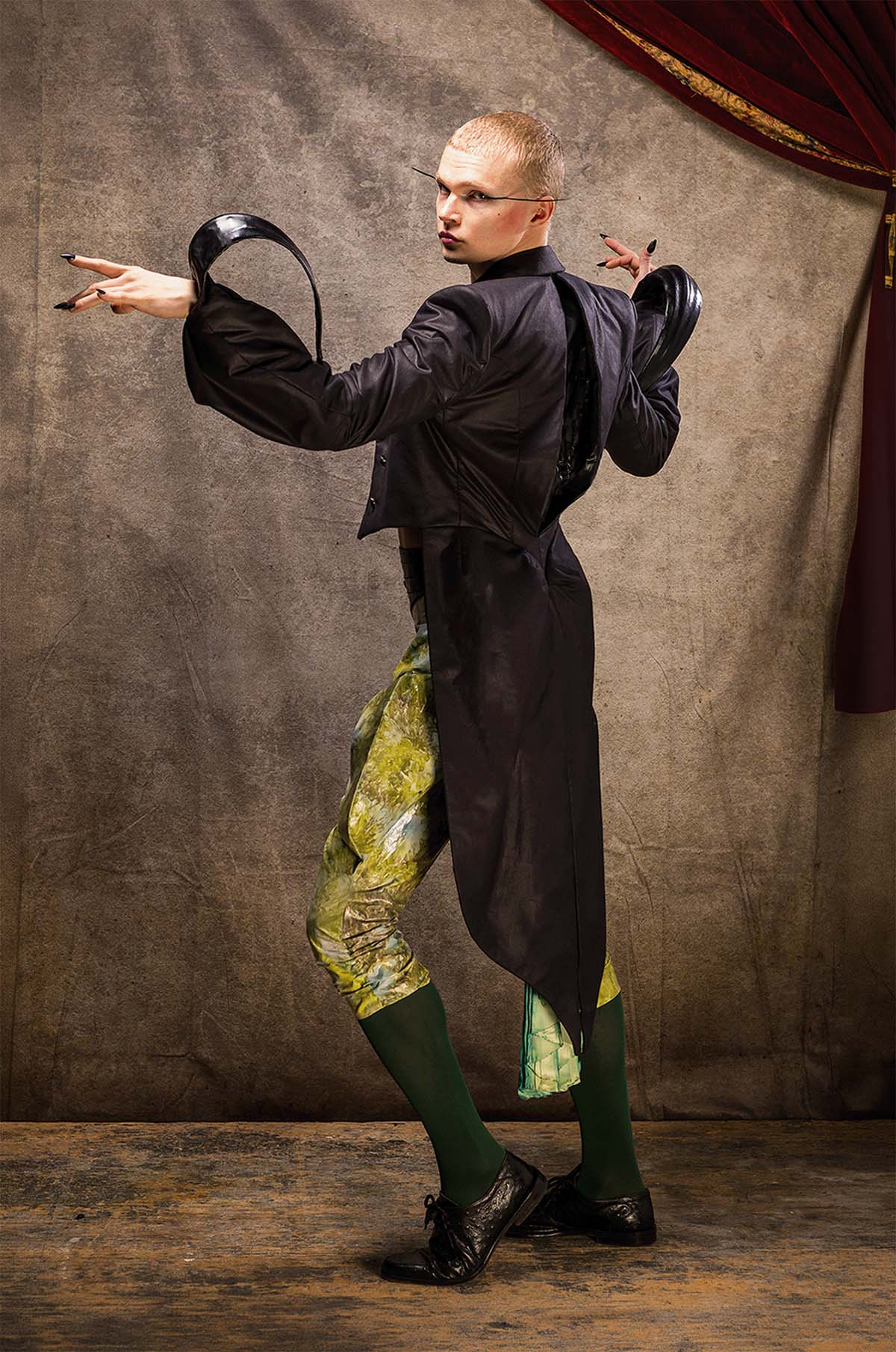 Costumed model seen from side back angle. Eerie design, Androgenous man in a state of Metamorphose, singular eyelashes extended beyond sides of face with harsh blushed cheeks. Clothed in a 1920's black Tailcoat with details in shiny black fabric over large cuffs which curl into a point arched up and over to elbow; a large gash-like pleat down centre back. Dyed Green mottled plastic wrapped Breeches scrunched at knee and protruded outwards and then curving in to the hip.
