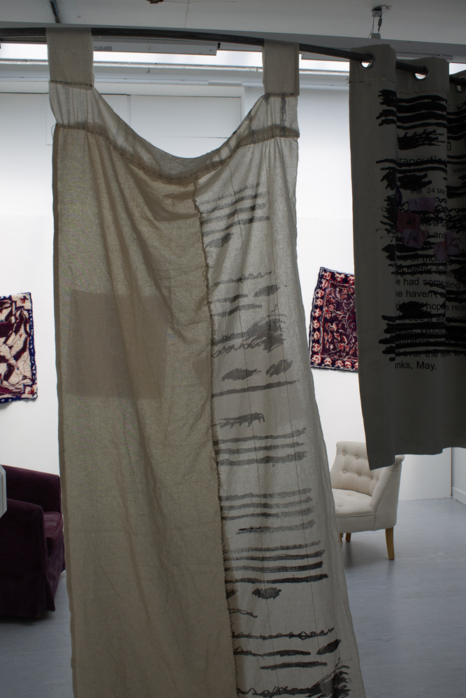 An image of hand-sewn curtains, printed with redactions, hung in front of two chairs in conversation and hand-made rugs hung on the wall  