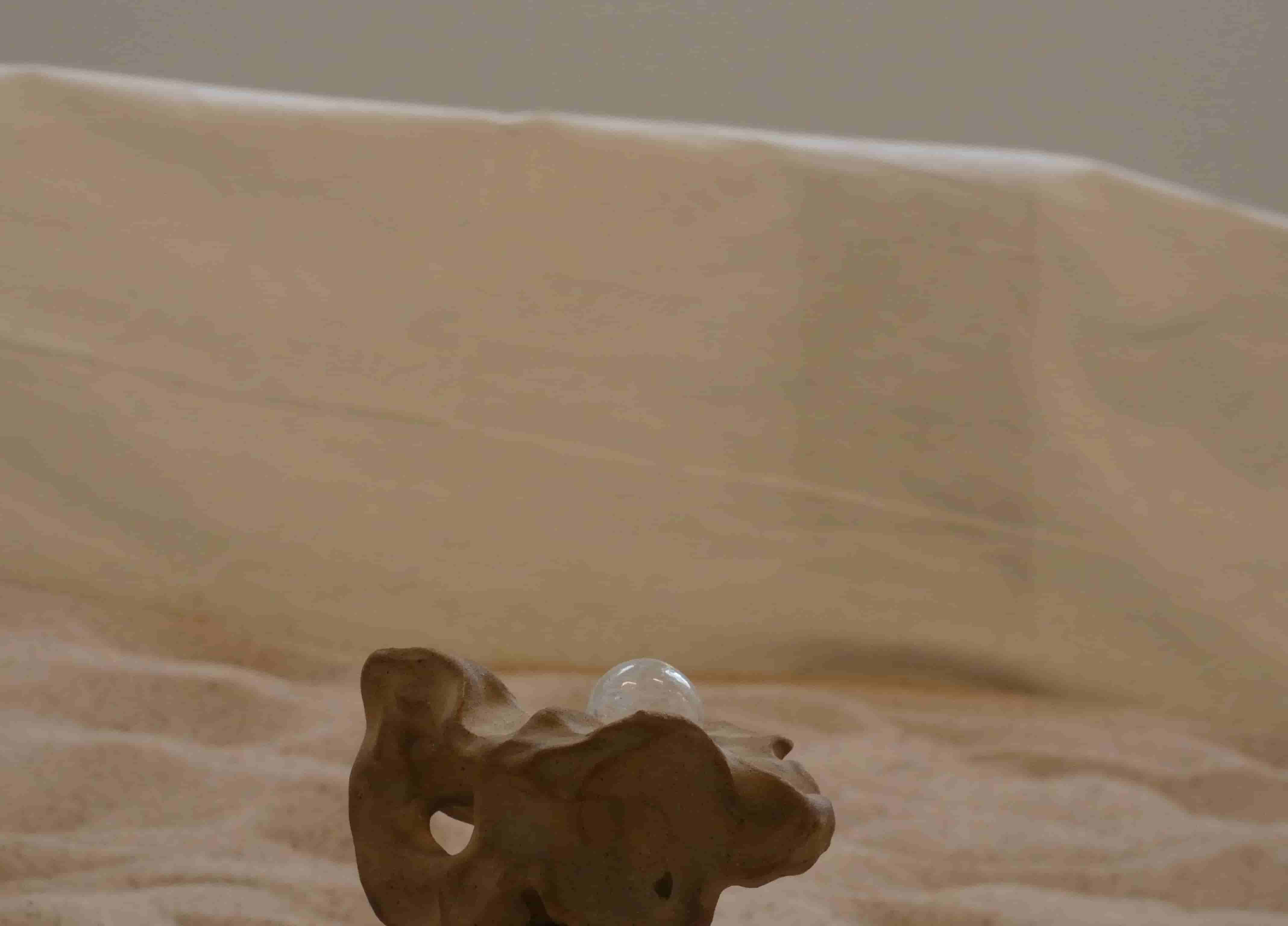 A close-up of part of an abstract ceramic sculpture with a clear glass marble balanced on it, sand and sheer fabric in background