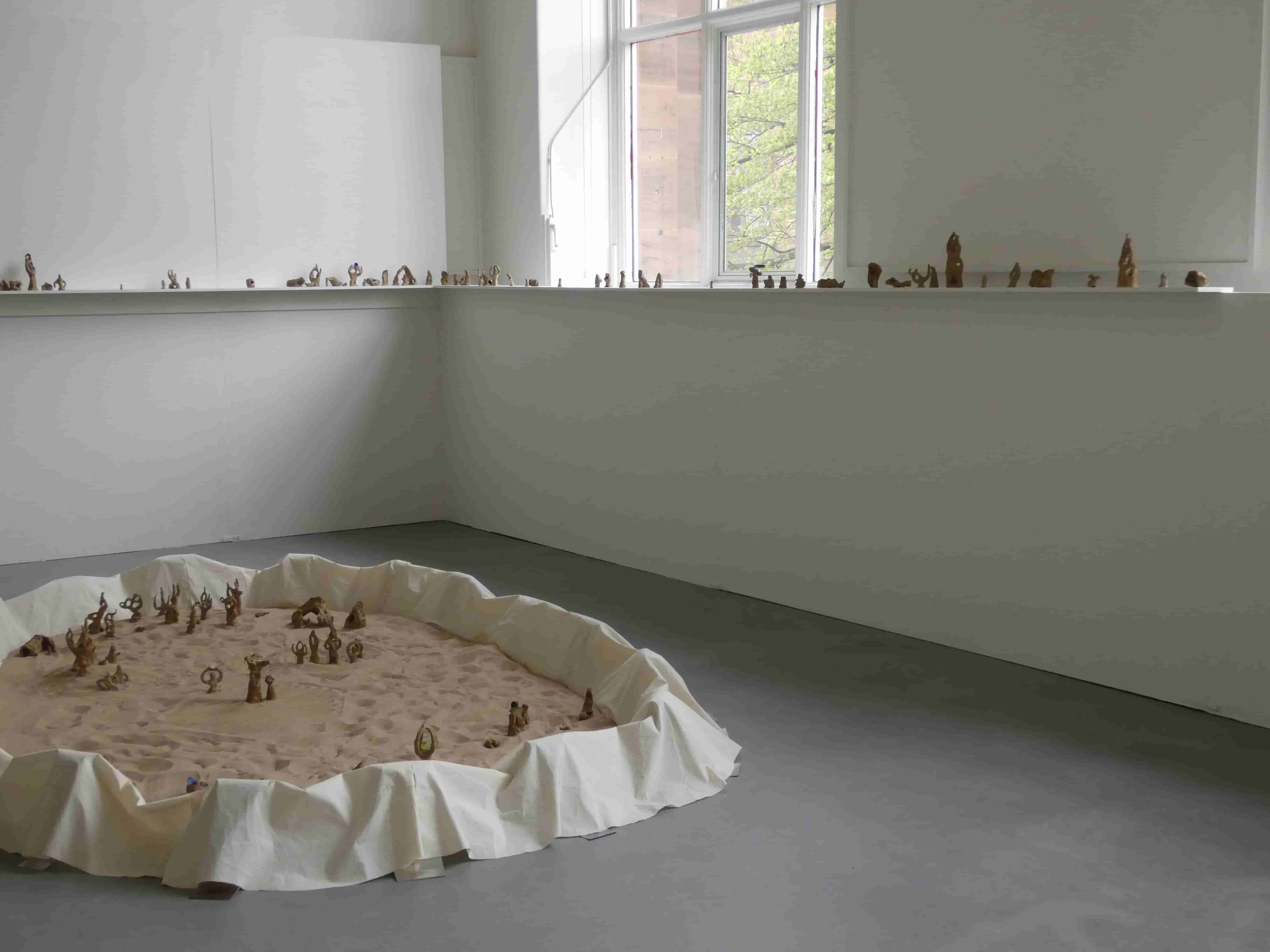 A wide view of the corner of a room, a single shelf displaying small sculptures runs along it, on the ground a round pit of sand held by fabric contains more sculptures