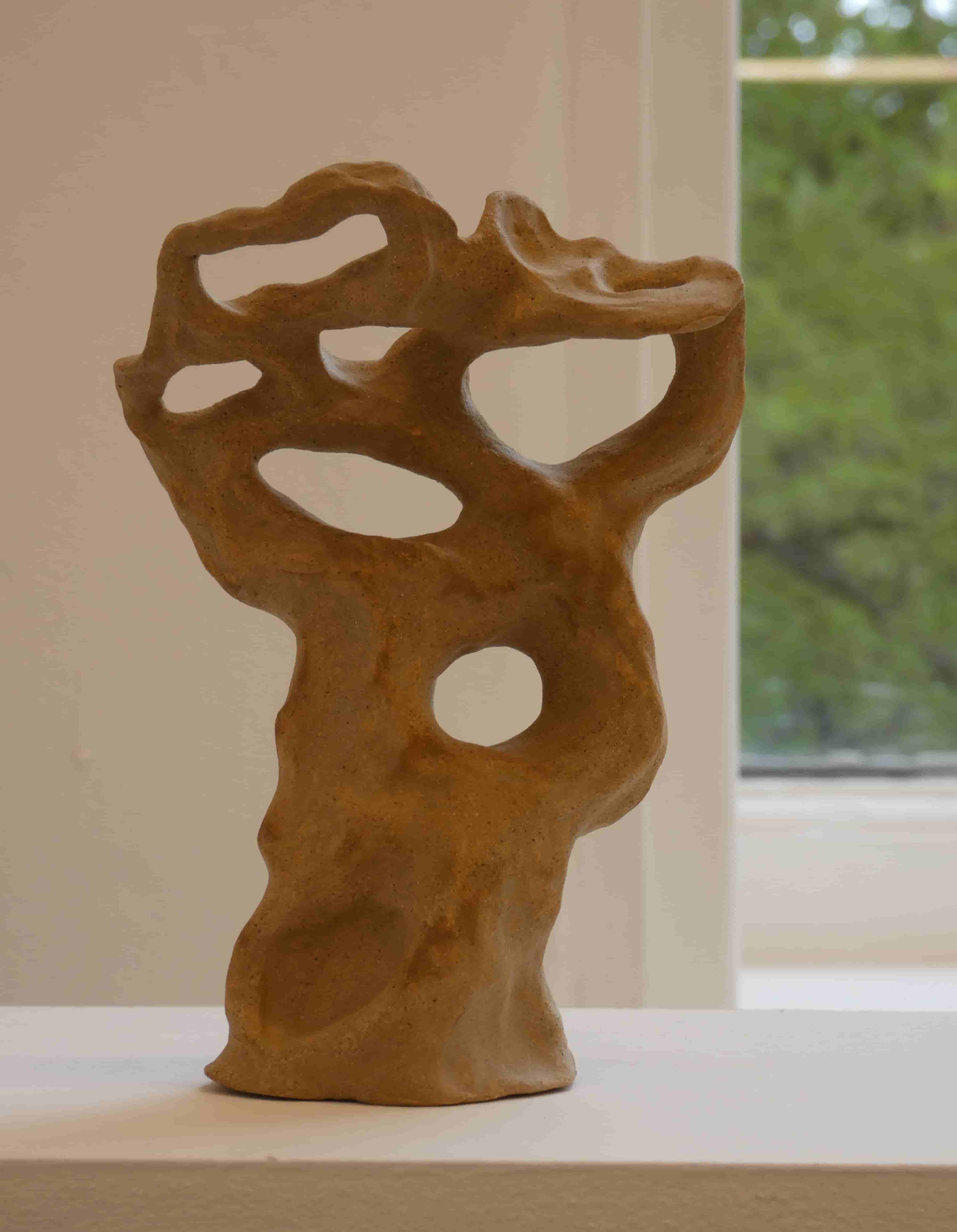 An abstract tree-like ceramic sculpture on a white shelf, white wall and part of a window with greenery in the background