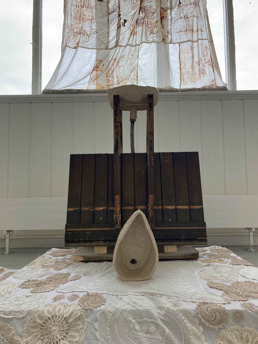 Image of sculptural instatllation including textiles hanging from a window, and a rug and wooden structure on the floor.