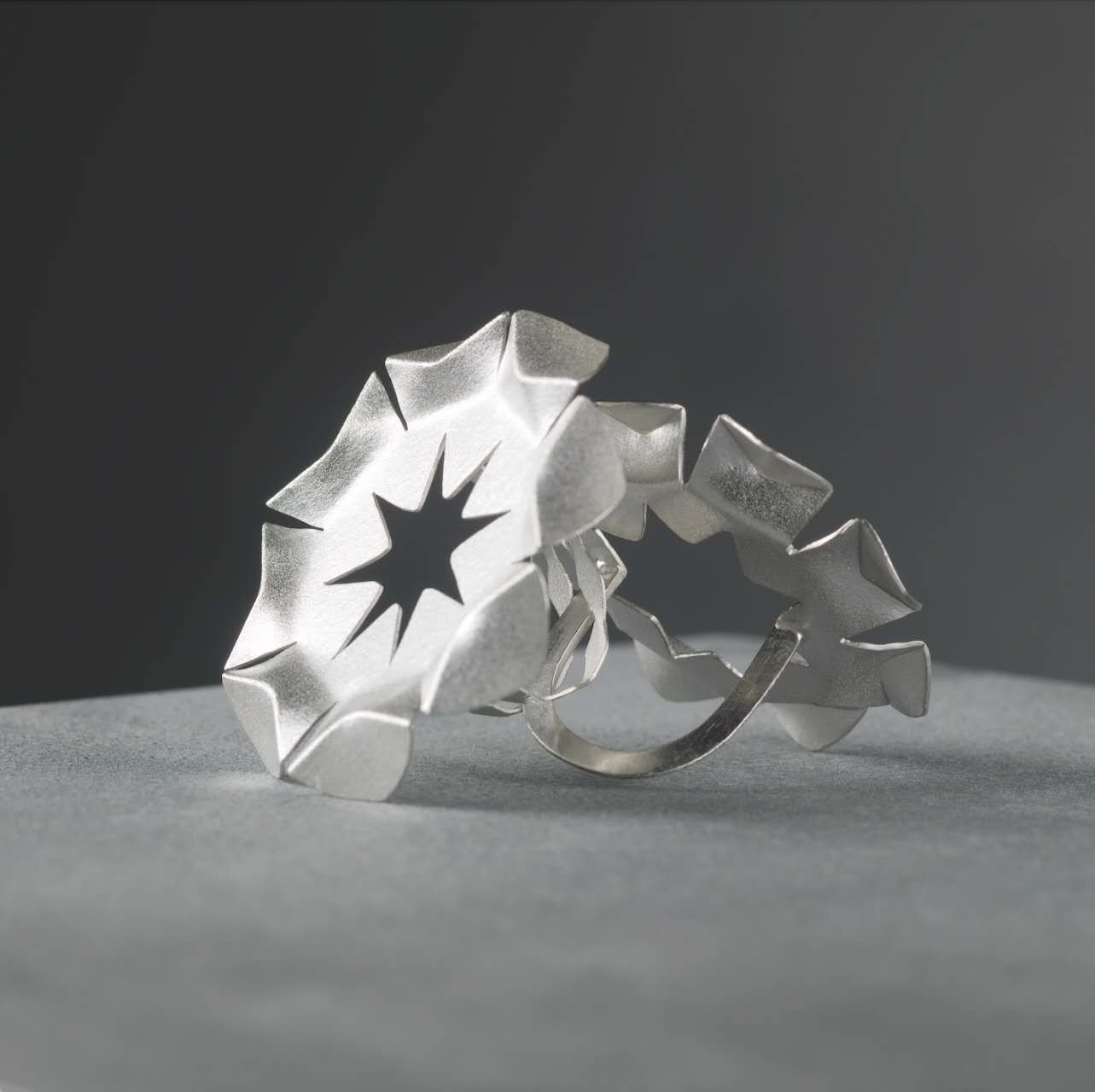 Geometric silver ring constructed from layered and folded metal forms, inspired by the repetitive arrangement systems of roof tiles and pinecones.
