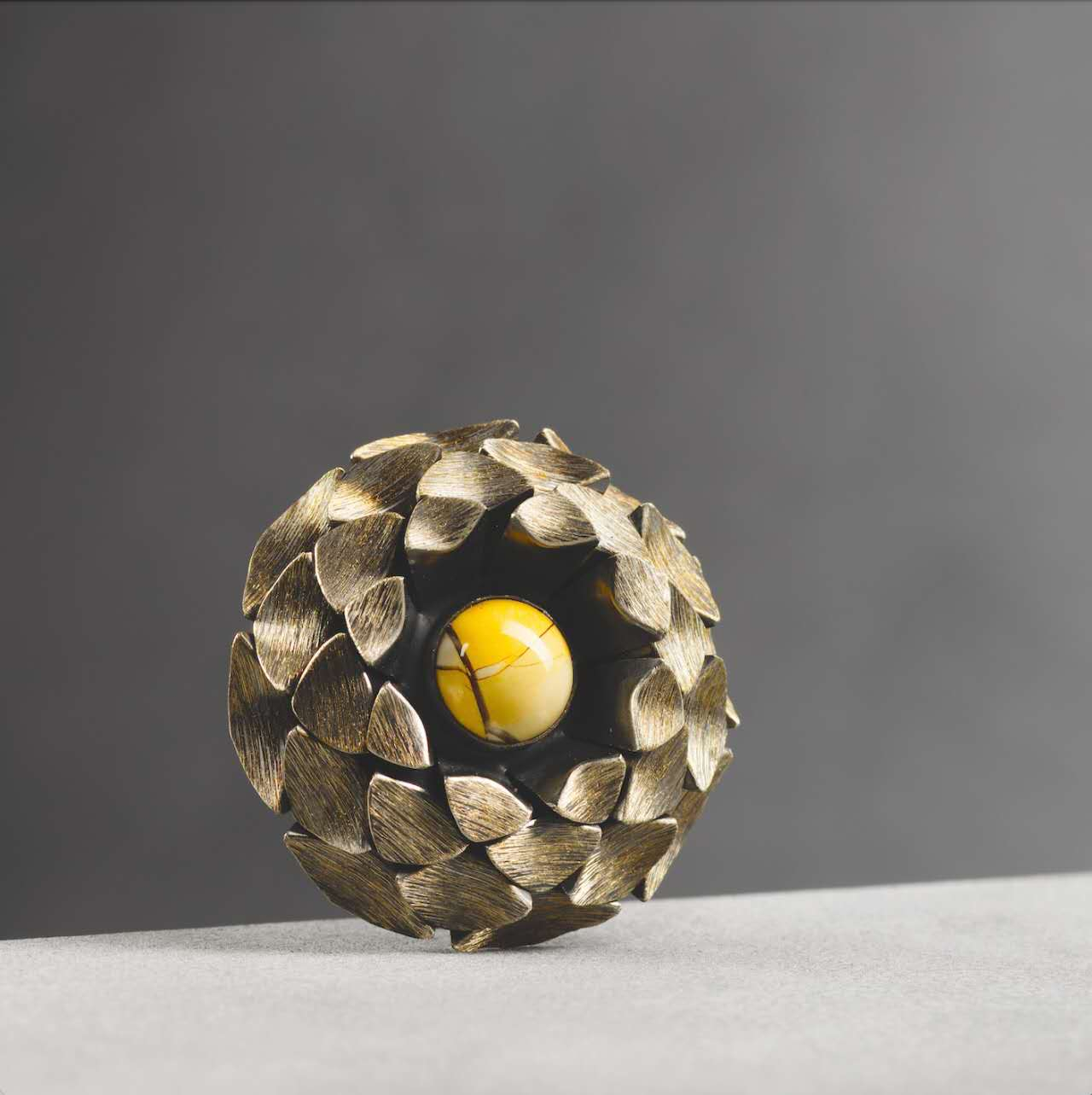 Circular layered brooch made of folded silver metal with gold elements arranged in a pinecone-like structure, centred with a polished brown jasper stone and featuring overlapping geometric forms inspired by natural growth patterns