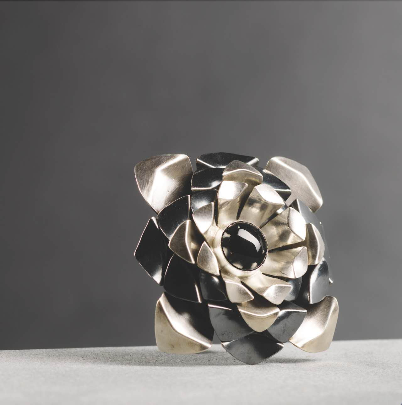 Square layered brooch made of folded silver metal arranged in a pinecone-like structure, centred with a polished obsidian stone and featuring overlapping geometric forms inspired by natural growth patterns