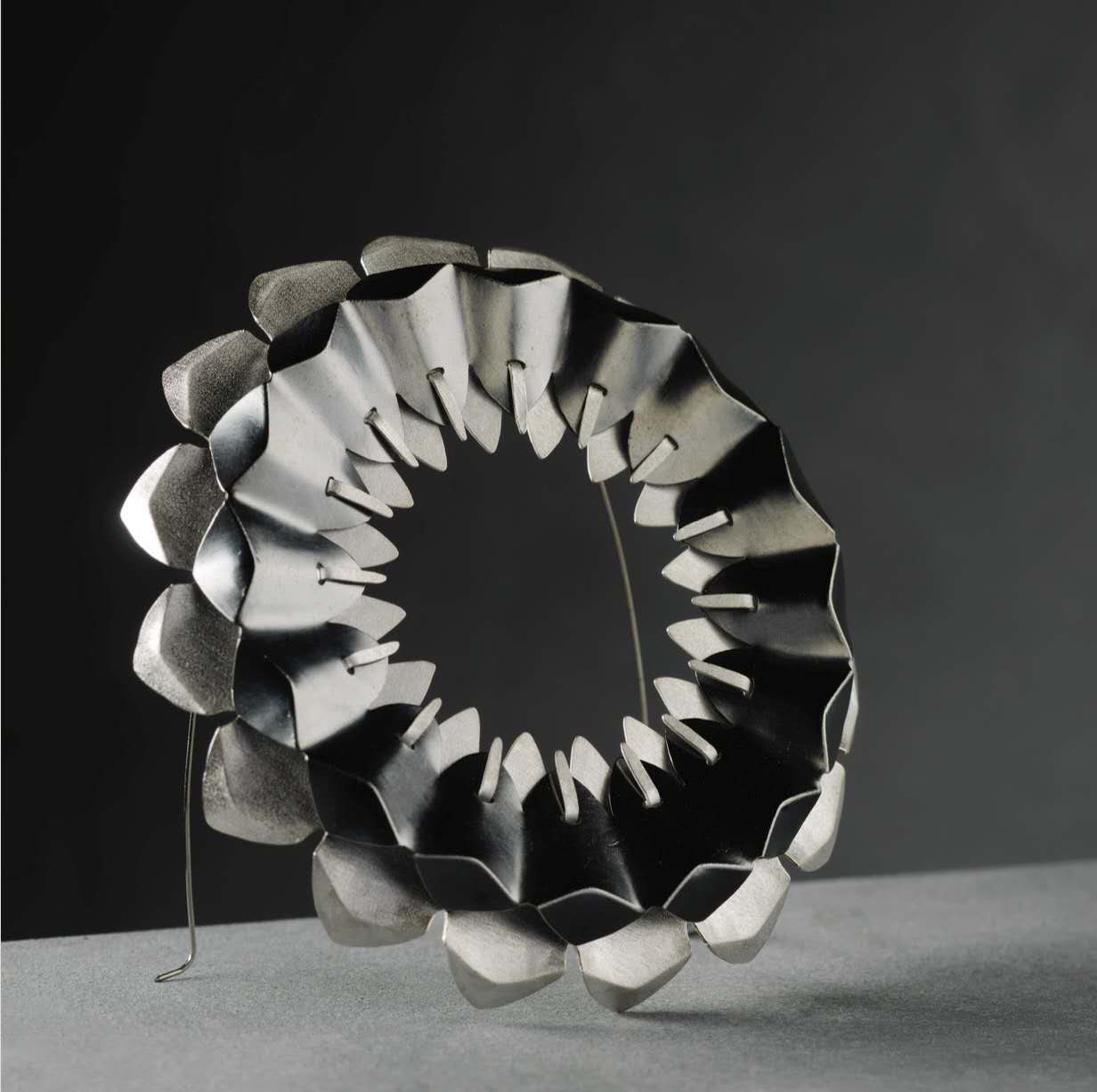 Large circular brooch composed of layered silver-toned metal forms in two contrasting colours, featuring an open hollow centre and overlapping folded elements inspired by pinecone and architectural structures