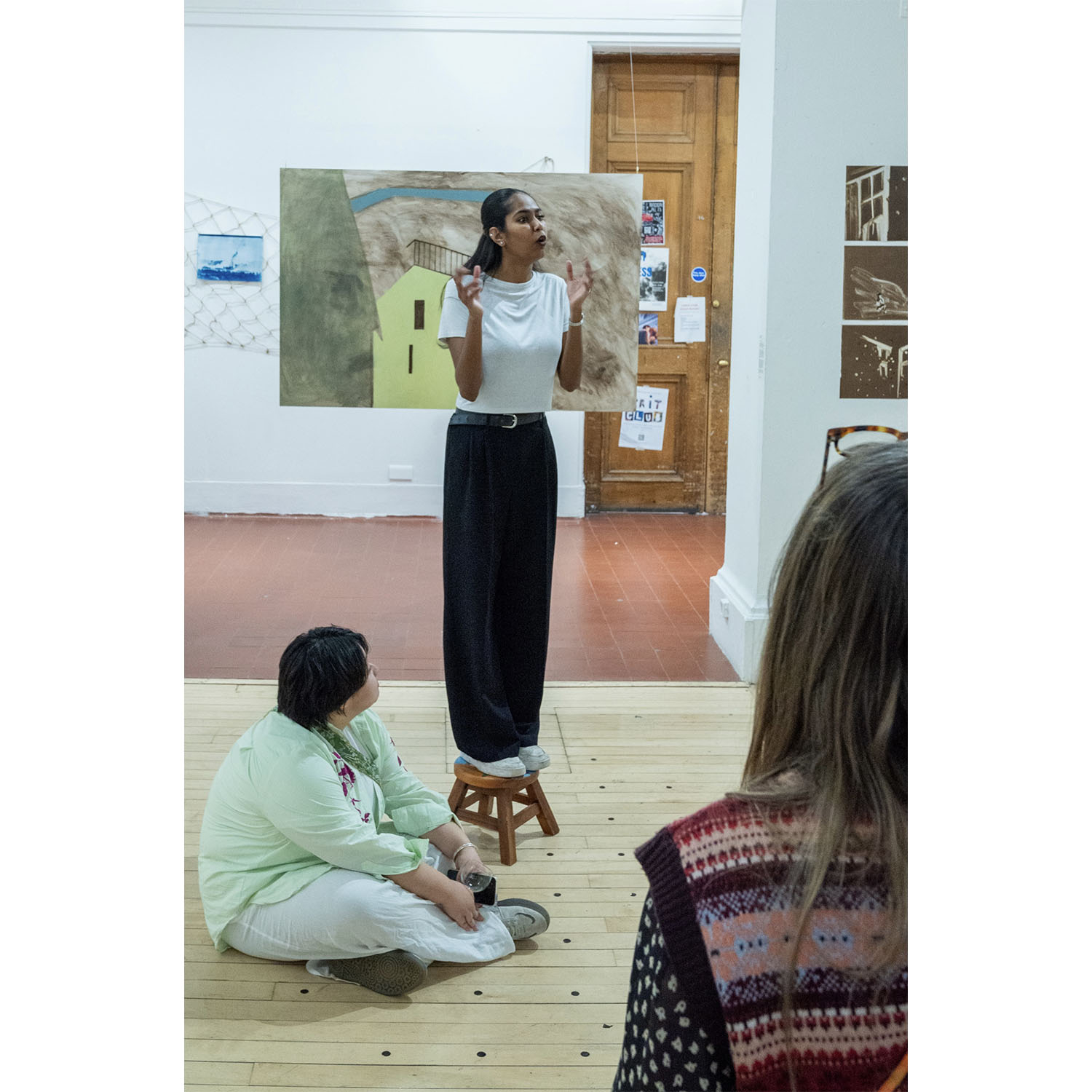     Full-body performance still of the artist standing on a wooden stool and gesturing while addressing an audience