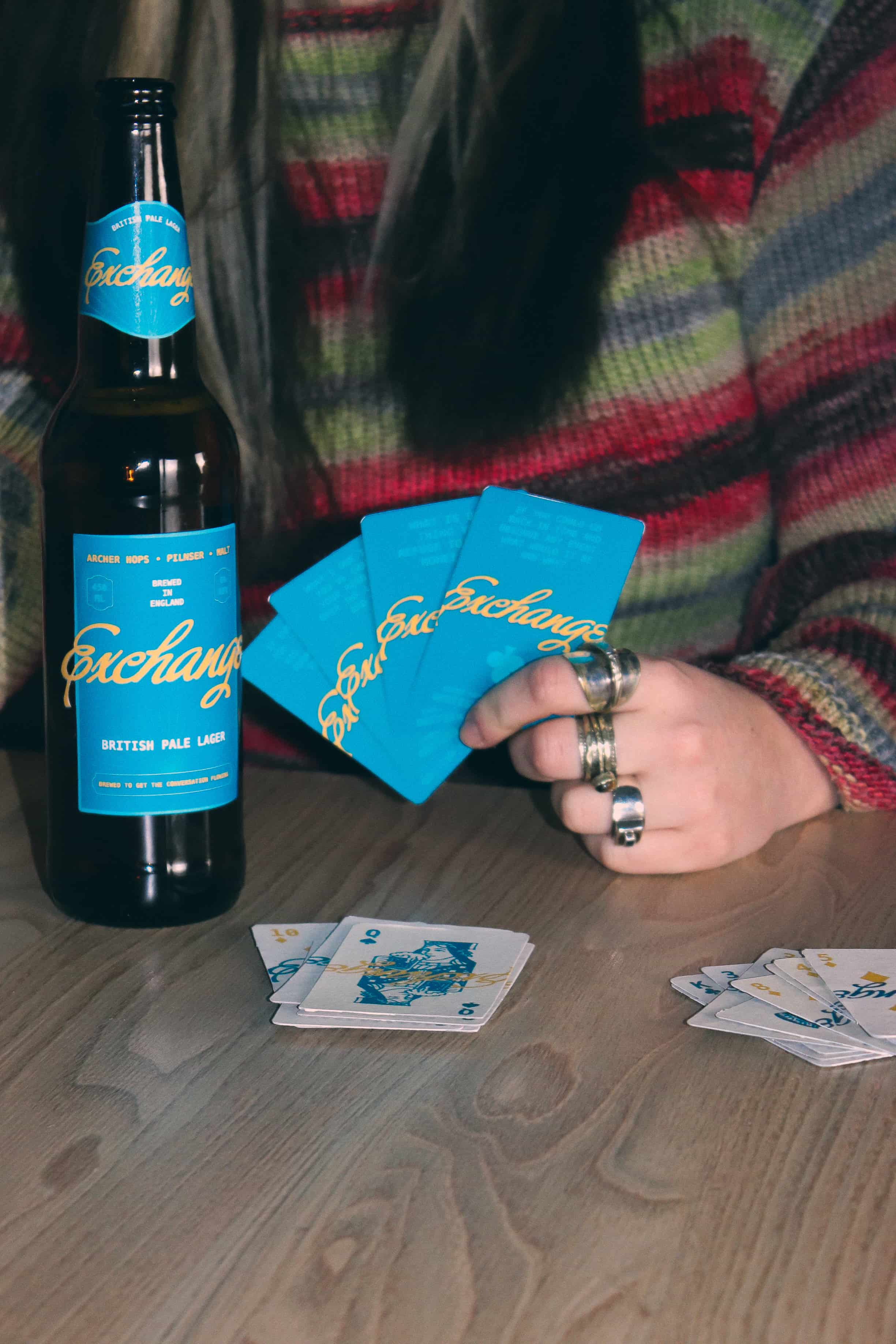    Close up of an ‘Exchange’ beer bottle on a table beside a person holding teal branded playing cards.