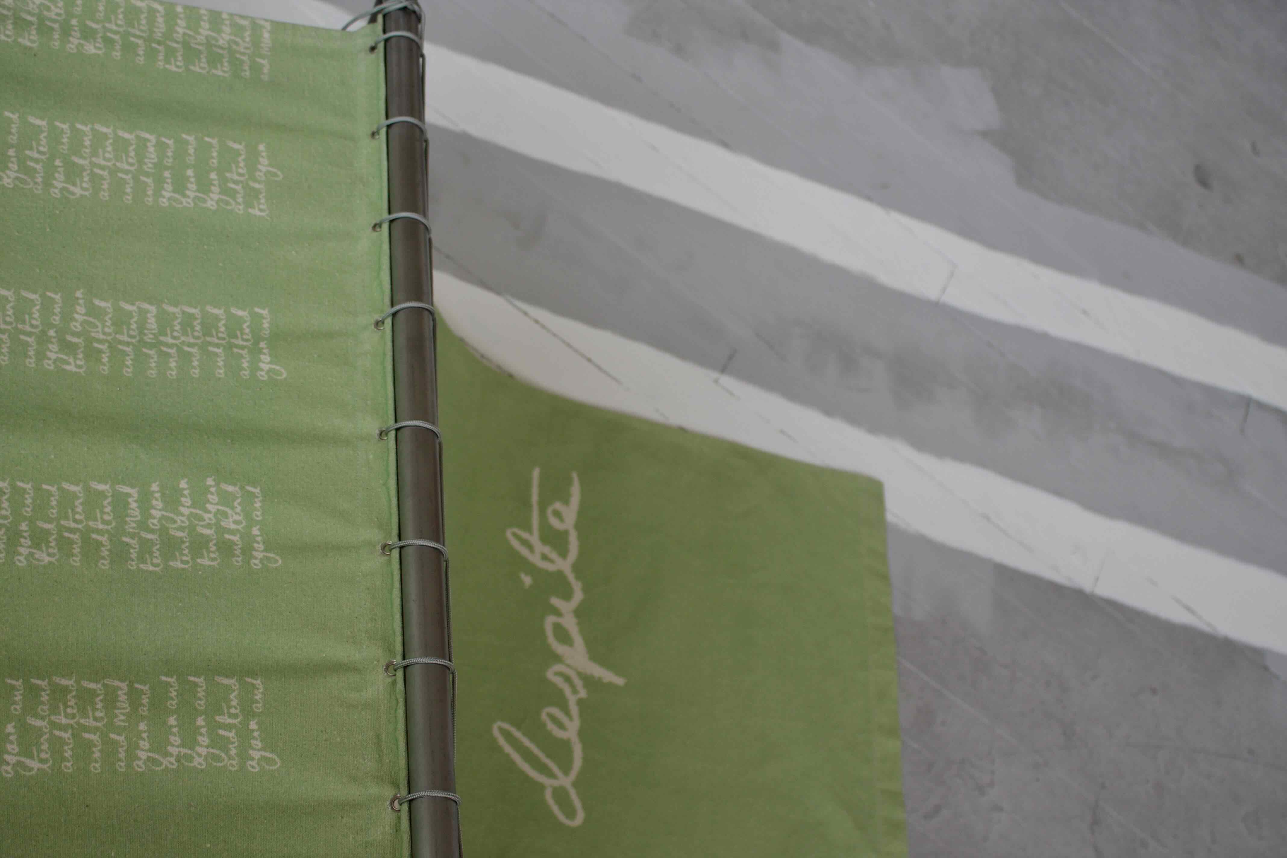 Details of green fabric with white text printed on - reading to tend again and again and again and tend again and again is to mend again and again and tend and mend again'. The fabric is attached to a steel pipe and suspended from a grey floor with two white lines painted on it.