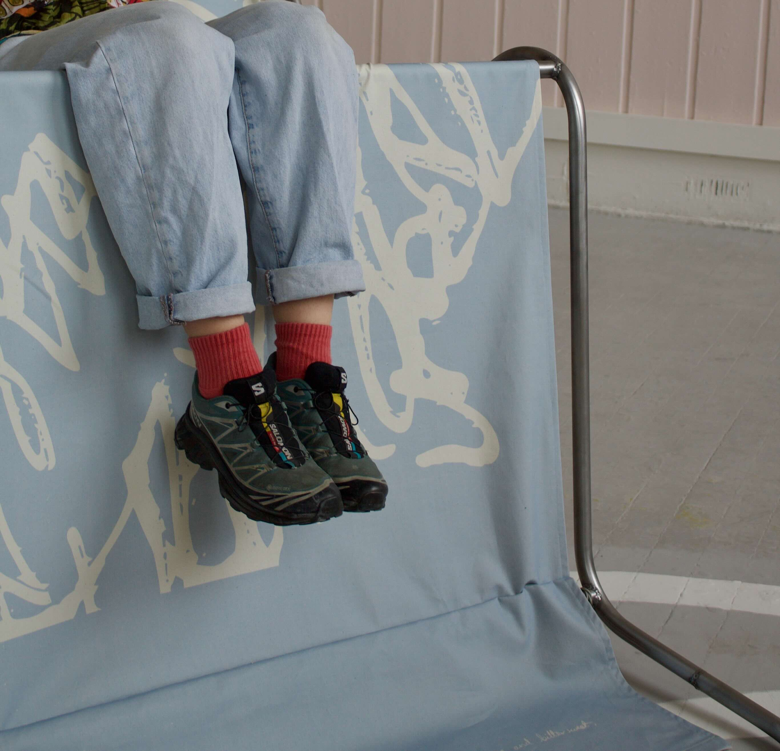 Legs overhanging a steel bar frame covered in light blue fabric with abstracted latter shapes printed on. Details of The Conversational Playground: To Twist, To Tend