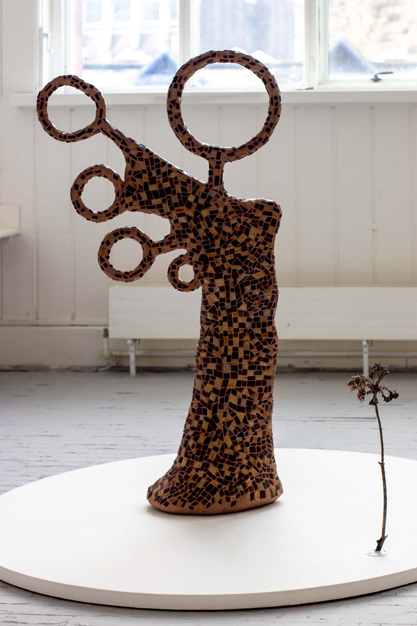 Abstract mosaic figure in brown ceramic tiles on a white round plinth, next to a bronze cast of hogweed.