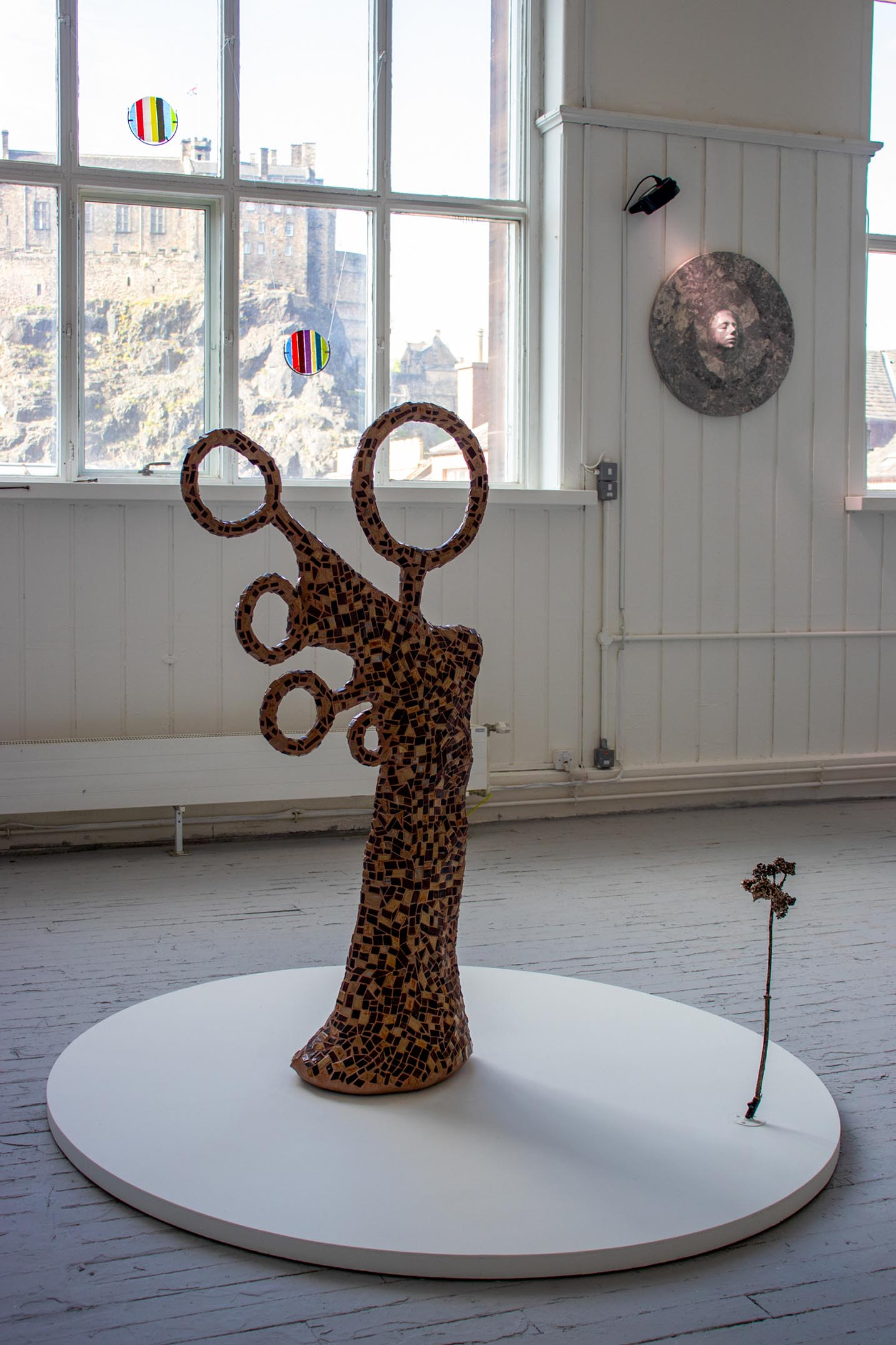 Installation view of an abstract mosaic figure in brown ceramic tiles on a white round plinth, beside a bronze cast of hogweed. Windows feature two circular pieces of fused glass.