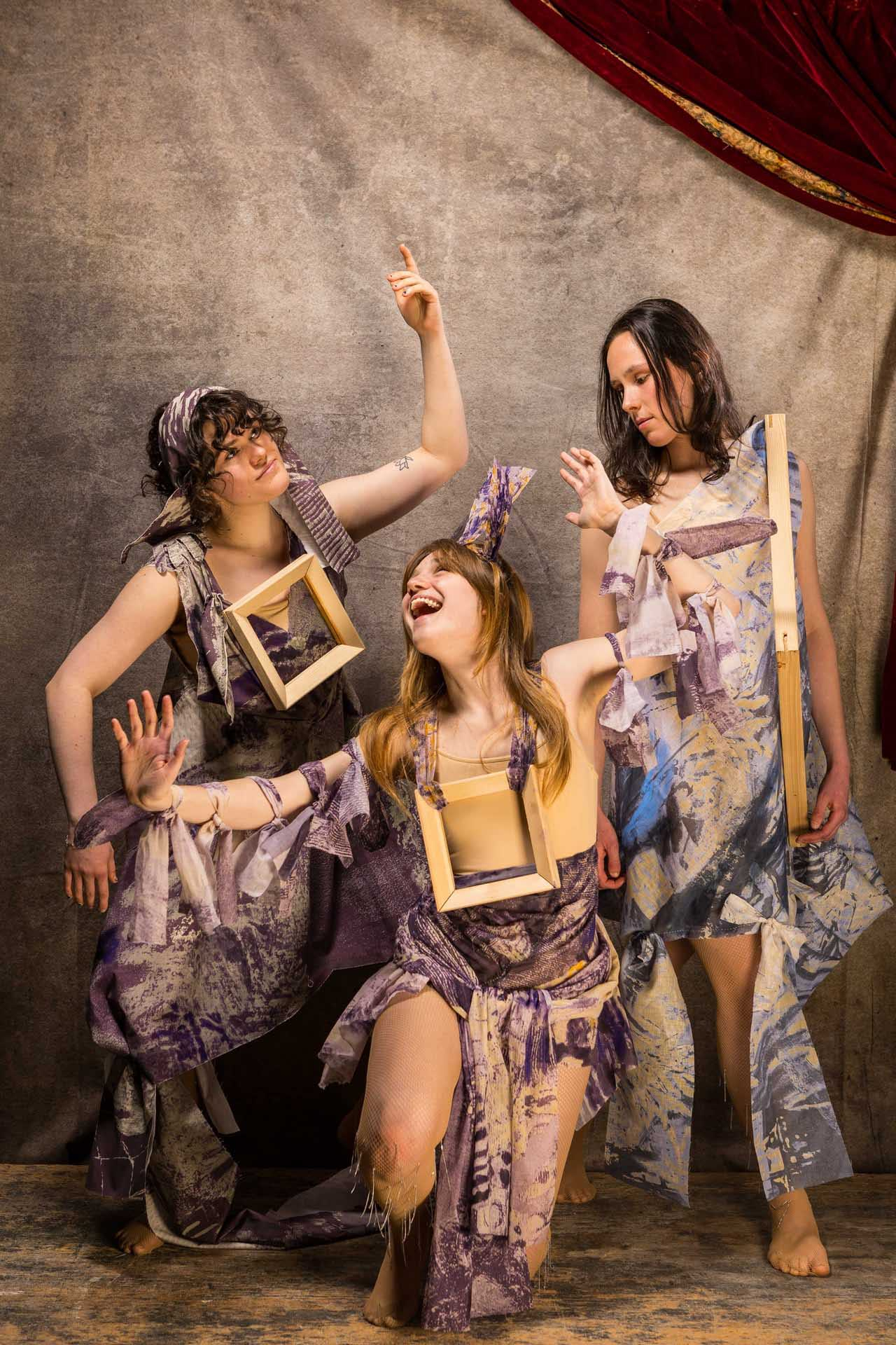 Three performers are wearing draped fabrics with painted prints in a theatrical setting.