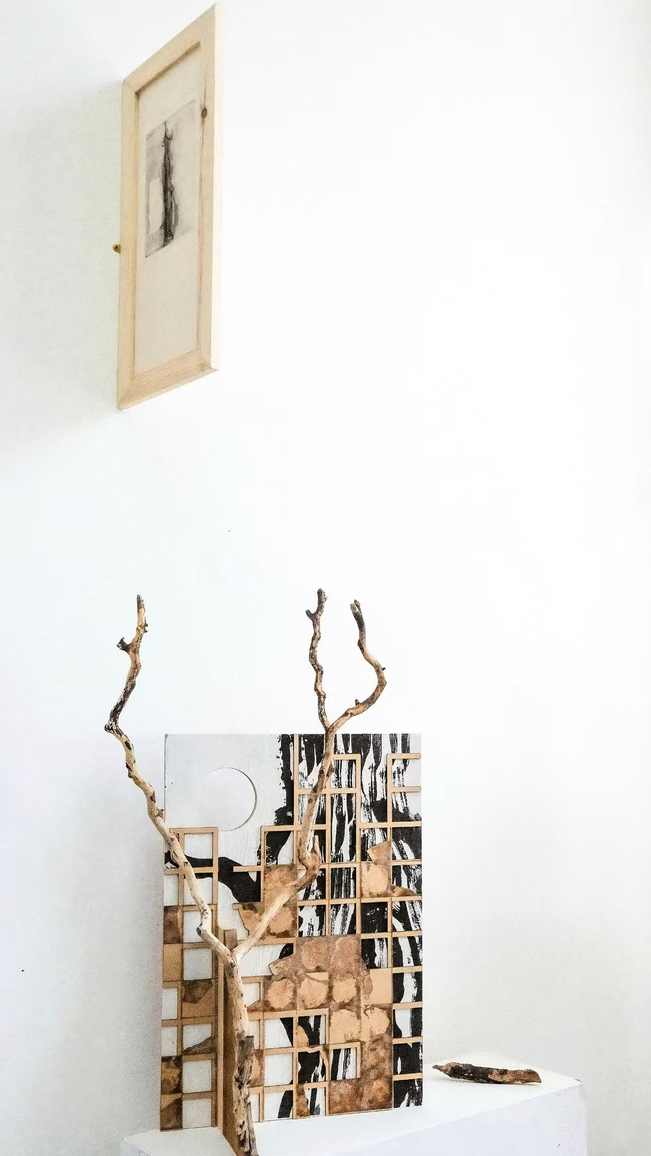 A mixed-media installation featuring a framed etching on the wall above a sculpture where a natural withered branch intersects a rigid wooden grid.