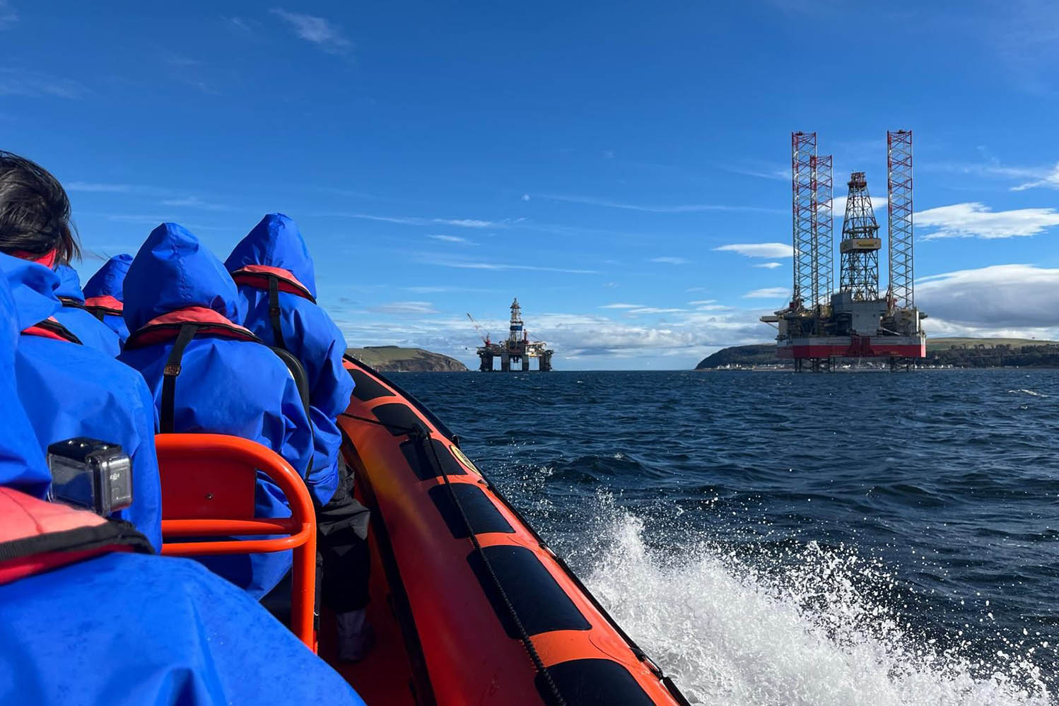 People wearing outdoor clothes have their backs to us and they are on a boat on the sea. They are speeding towards two oil rigs in the distance. The sky is blue.