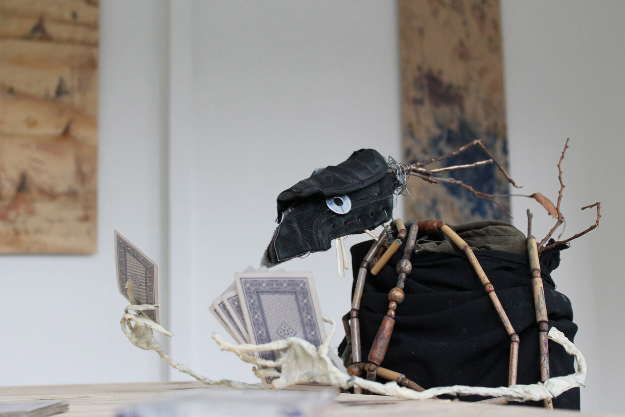 A curious puppet-like creature made of black fabric is holding playing cards in its skeletal hands. Its eyes are made of metal washers..